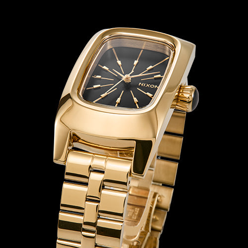 A close-up of a gold Nixon watch with a rectangular black dial and gold hour markers, set against a black background.