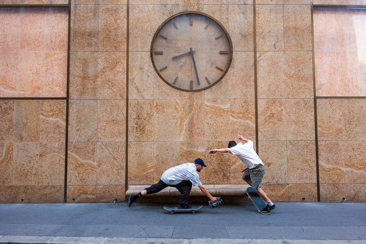 A skateboarder does a trick on a bench under a large clock while a videographer films